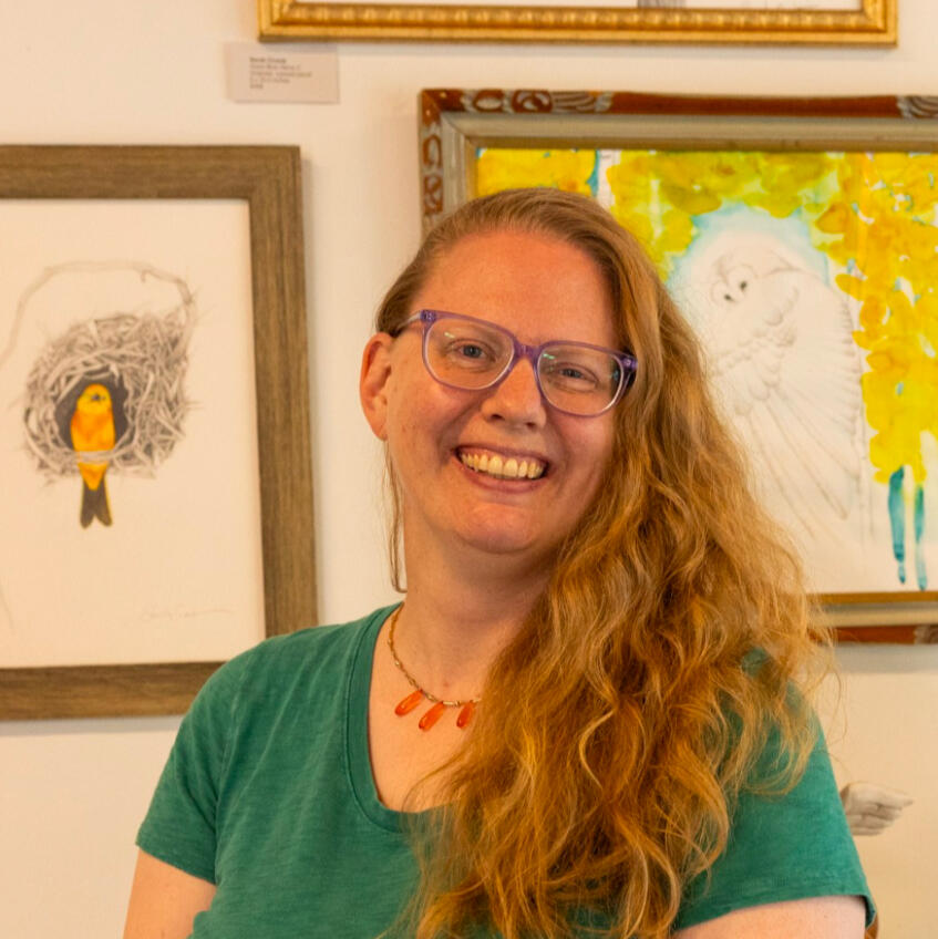 Sarah and some of her work Adult blonde lady smiling in front of silver art on a wall.
